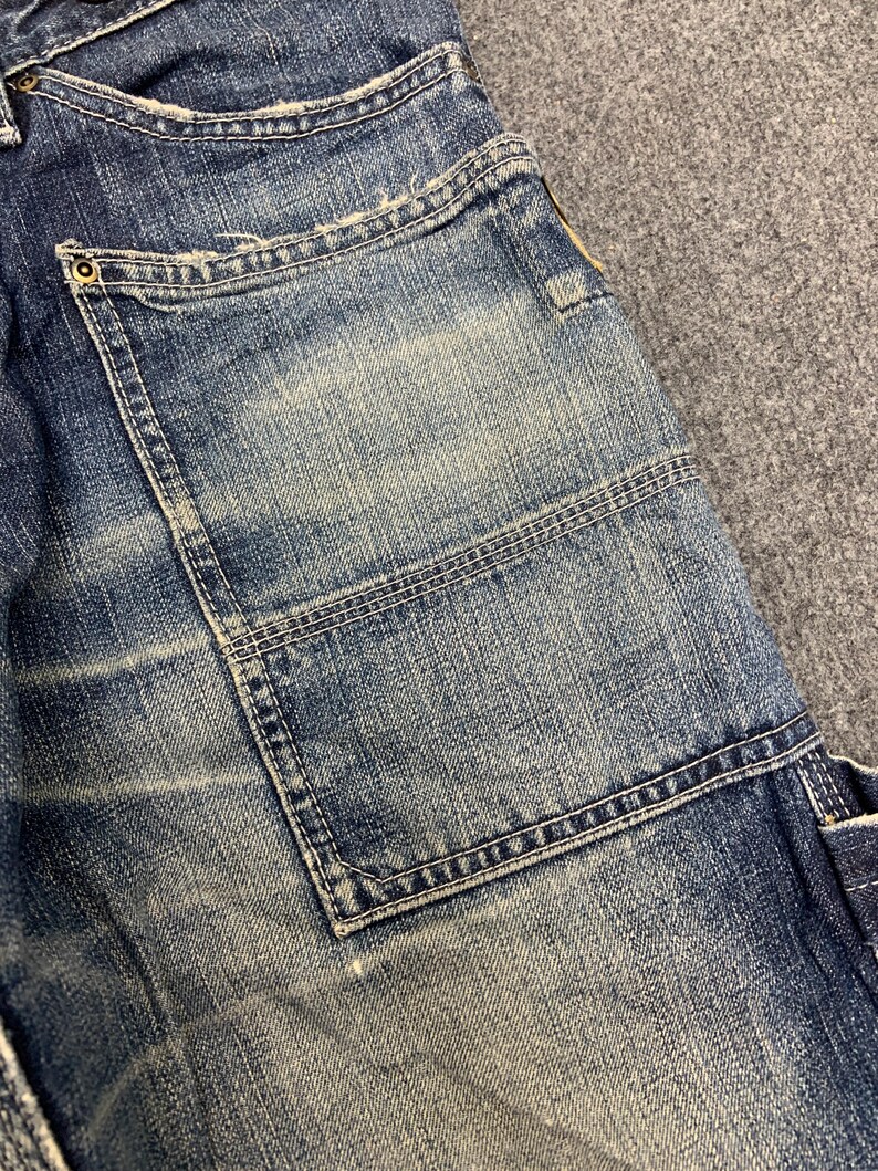 Puede incluir: Primer plano de una secci&oacute;n de vaqueros azules. Los vaqueros tienen un bolsillo grande y cuadrado con costuras reforzadas. El tejido vaquero muestra signos de desgaste y decoloraci&oacute;n, con zonas m&aacute;s claras que contrastan con el tejido azul m&aacute;s oscuro. Se ve un peque&ntilde;o remache met&aacute;lico.