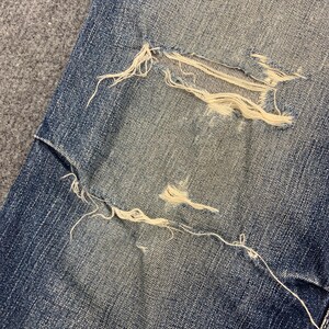 Puede incluir: Primer plano de vaqueros de mezclilla azul desgastados con una gran secci&oacute;n rasgada que expone hilos blancos. La mezclilla tiene una apariencia descolorida, con desgaste visible. Los bordes deshilachados y los hilos expuestos a&ntilde;aden a la est&eacute;tica vintage.