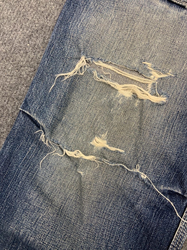 Puede incluir: Primer plano de vaqueros de mezclilla azul desgastados con una gran secci&oacute;n rasgada que expone hilos blancos. La mezclilla tiene una apariencia descolorida, con desgaste visible. Los bordes deshilachados y los hilos expuestos a&ntilde;aden a la est&eacute;tica vintage.