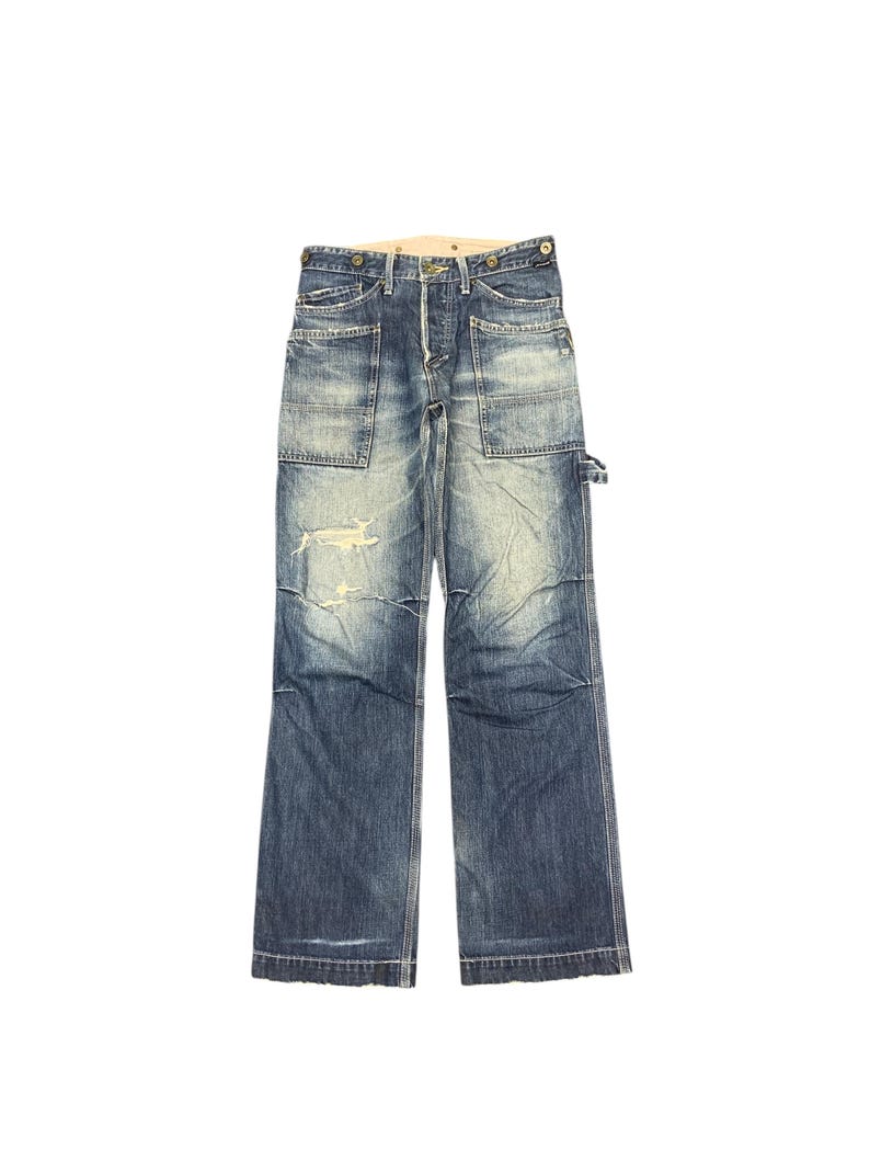 Puede incluir: Pantalones de trabajo vaqueros azules descoloridos con m&uacute;ltiples bolsillos y detalles desgastados. Los vaqueros de estilo vintage tienen bragueta con botones y un corte relajado. Los pantalones tienen un lavado claro con desgaste visible, d&aacute;ndoles un aspecto usado.