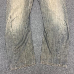 May include: Faded denim jeans with a straight leg cut. The jeans have a worn, distressed appearance with lighter areas on the thighs and knees. The fabric has a textured look.