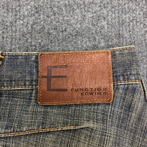 May include: Close-up of blue denim jeans with a brown leather patch. The patch features the brand name "FUNCTION EDWIN" and a large "E" design. The jeans have visible stitching and a textured appearance.