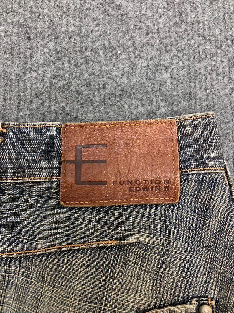 May include: Close-up of blue denim jeans with a brown leather patch. The patch features the brand name "FUNCTION EDWIN" and a large "E" design. The jeans have visible stitching and a textured appearance.