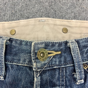 Puede incluir: Primer plano de la cintura de unos vaqueros azules. Los vaqueros tienen un bot&oacute;n con el texto "AVIREX U.S.A." y una cinturilla interior beige. Los vaqueros tienen costuras visibles y remaches met&aacute;licos.