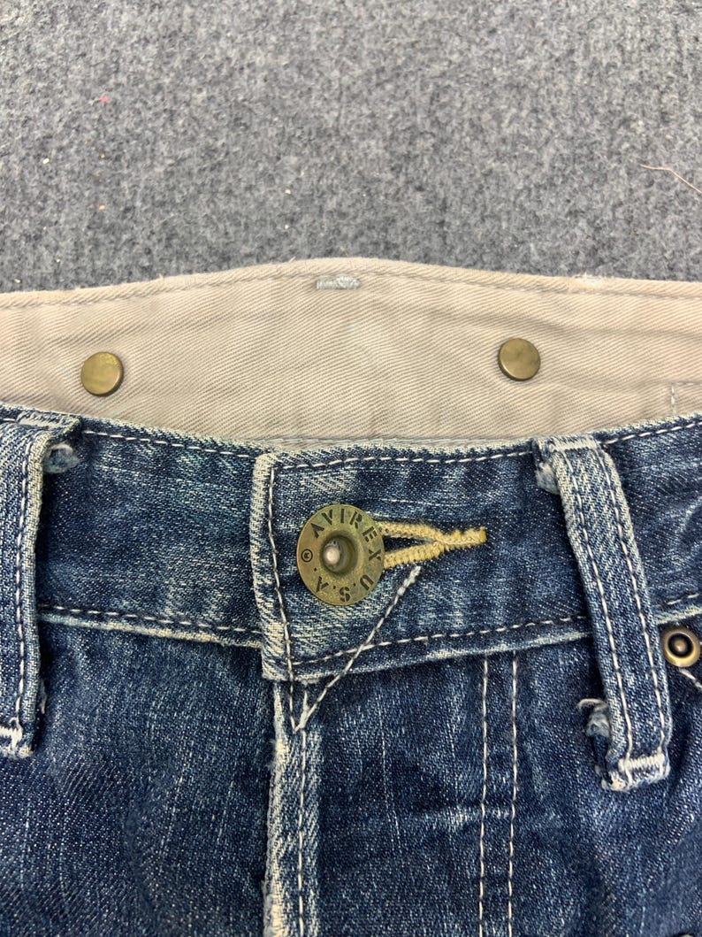 Puede incluir: Primer plano de la cintura de unos vaqueros azules. Los vaqueros tienen un bot&oacute;n con el texto "AVIREX U.S.A." y una cinturilla interior beige. Los vaqueros tienen costuras visibles y remaches met&aacute;licos.