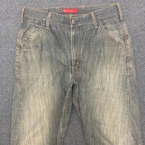 May include: Faded blue denim jeans with a classic five-pocket design. The jeans have a button and zipper closure, belt loops, and a subtle faded wash effect. The brand label is visible on the waistband.