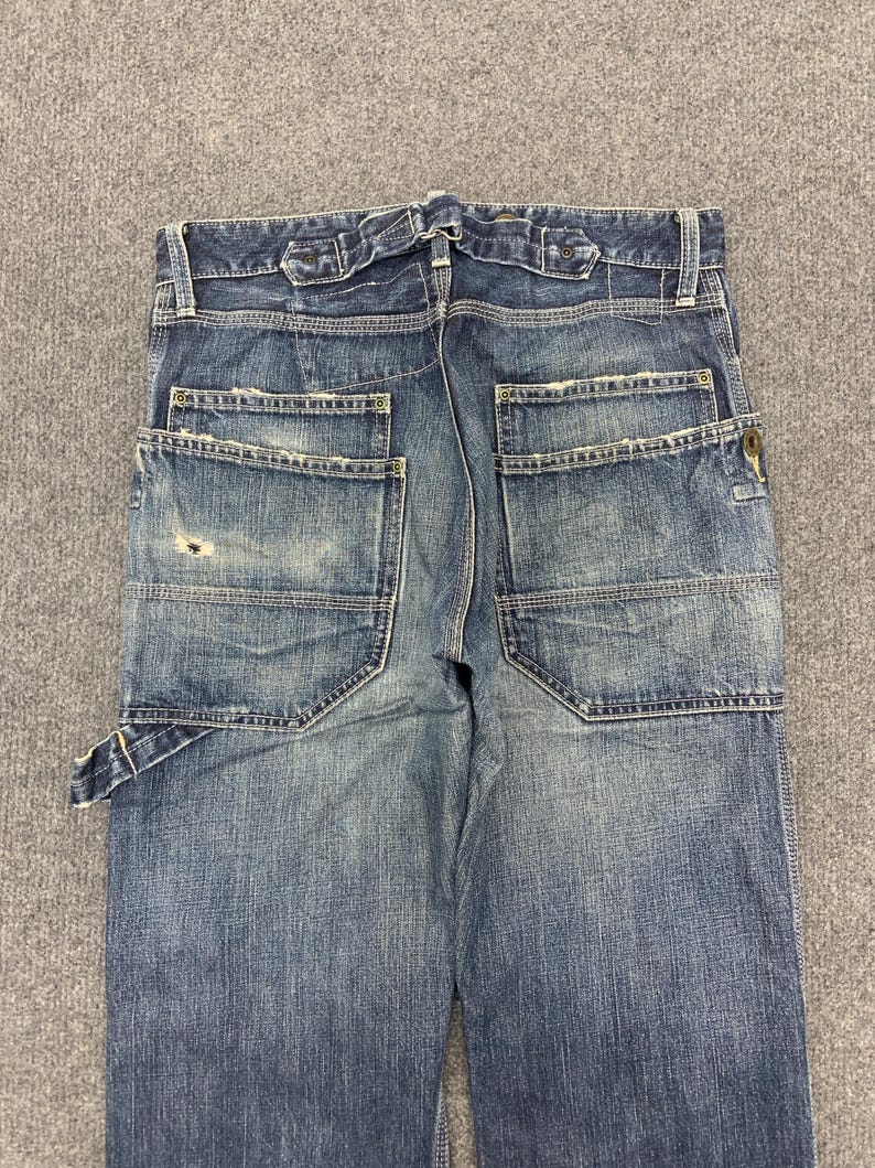 Puede incluir: Vaqueros de mezclilla azul con un aspecto desgastado, con m&uacute;ltiples bolsillos y un dise&ntilde;o de estilo carpintero. Los vaqueros tienen un lavado descolorido y detalles de costura visibles, con cierre de bot&oacute;n y cremallera.