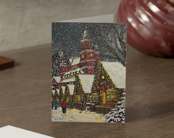 May include: A folded greeting card with a winter scene of a village with snow-covered buildings and people walking. A second, blank card is in front. The cards are on a wooden surface, with a decorative vase and mirror in the background.
