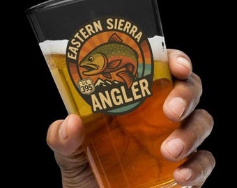 May include: A pint glass filled with beer, held by a hand. The glass features a circular logo with the words "Eastern Sierra Angler" and a fish illustration. The logo also includes "US 395".