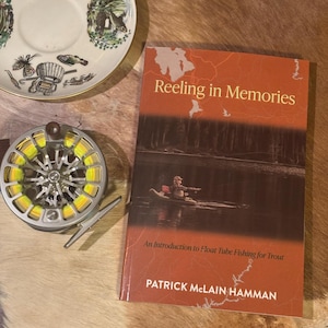 May include: A fly fishing reel with yellow line, a book titled "Reeling in Memories," and a decorative plate with fishing-themed illustrations. The book cover features an angler in a float tube. The reel is silver and yellow.