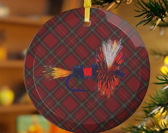May include: Round Christmas ornament with a red plaid pattern background. The ornament features a colorful illustration of a fishing fly. The fly has orange, yellow, white, and red details. A gold ribbon is attached for hanging.