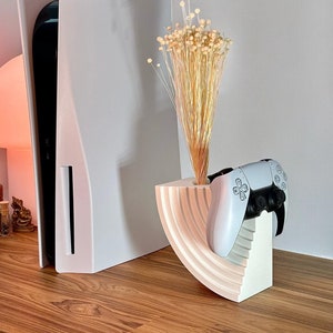 May include: White PlayStation 5 console with a white controller stand. The stand has a curved design and holds a white PlayStation 5 controller. Dried flowers are in the stand.