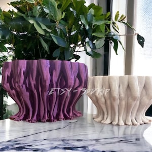 May include: Two 3D printed planters shaped like a group of legs. The planter on the left is a deep purple color and the planter on the right is a light beige color. Both planters are sitting on a marble surface.