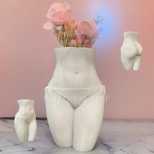 May include: White ceramic vase shaped like a female torso with a bouquet of pink flowers. The vase is sitting on a white marble surface.