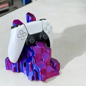 May include: A white video game controller sits on a purple and blue crystal-shaped stand. The stand is made of 3D printed plastic.