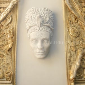 May include: A white marble sculpture of a serene face with ornate headwear, including a cobra. The sculpture is mounted on a light-colored wall, flanked by intricately carved golden architectural elements. The word "GALLERY" is partially visible.