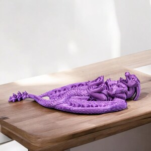 May include: A purple 3D printed dragon figurine with detailed scales and wings. The dragon is curled up in a sleeping position.