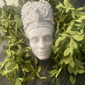 May include: A white, carved bust of a woman's face, adorned with an ornate headdress. The bust is surrounded by green leafy vines, creating a naturalistic frame. The sculpture has detailed facial features and a serene expression.