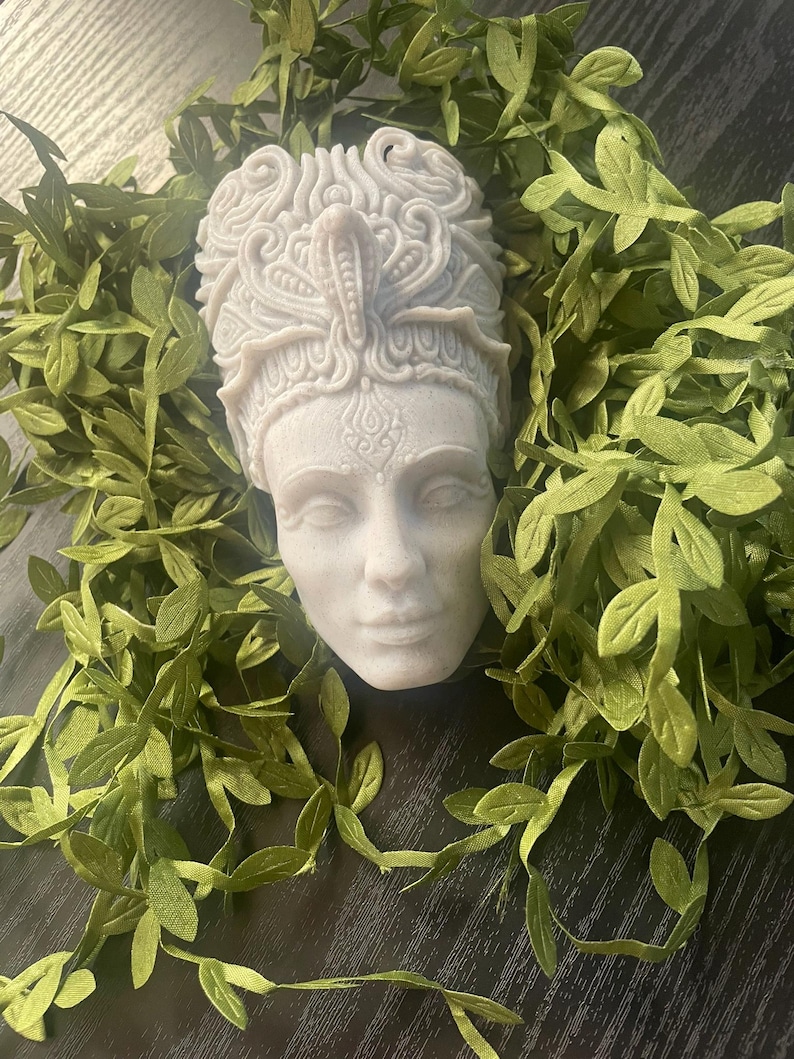 May include: A white, carved bust of a woman's face, adorned with an ornate headdress. The bust is surrounded by green leafy vines, creating a naturalistic frame. The sculpture has detailed facial features and a serene expression.