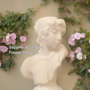 May include: White ceramic bust of a woman with a flower pot vase in the top of her head. The bust is against a background of pink flowers and green leaves. The text "Sappho of Lesbos Flower Pot Vase" is visible in the image.