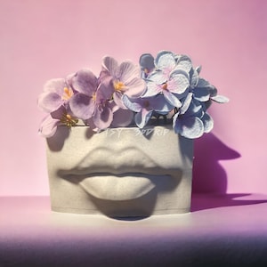 May include: A white ceramic planter shaped like a pair of lips with a bouquet of purple and blue flowers.