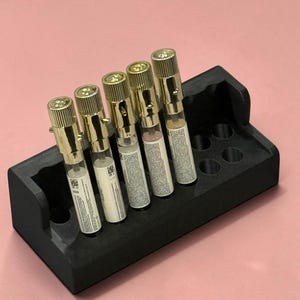 May include: Five small glass perfume vials with gold-toned metallic caps are arranged in a row within a black rectangular foam holder. Each vial features a white label with printed text and a small QR code, standing upright in the foam base.