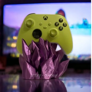 May include: A purple crystal-shaped stand holds a lime green Xbox controller. The controller has a black Xbox logo and black buttons.