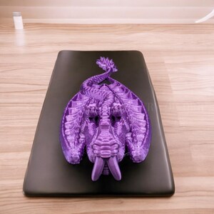 May include: A purple 3D printed dragon figurine with a curled tail and an open mouth, lying on a black surface.
