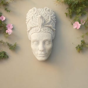 May include: A white, sculpted face with ornate headwear, mounted on a beige wall. The headpiece features a cobra and intricate patterns. Pink flowers and green vines frame the sculpture, creating a serene, artistic display.