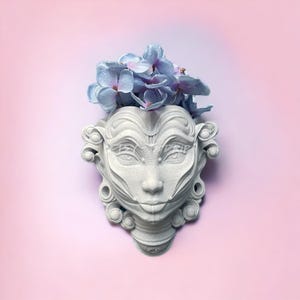May include: A white ceramic planter shaped like a face with intricate details, holding a bouquet of light blue flowers.