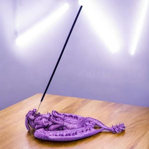 May include: Purple dragon incense holder with a black incense stick burning. The dragon is lying on its side with its wings spread out.