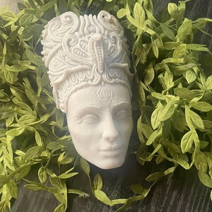 May include: A detailed, off-white sculpture of a regal female bust, adorned with an elaborate headdress featuring a cobra. The bust is surrounded by green leafy vines, creating a naturalistic contrast. The sculpture's face has a serene expression.
