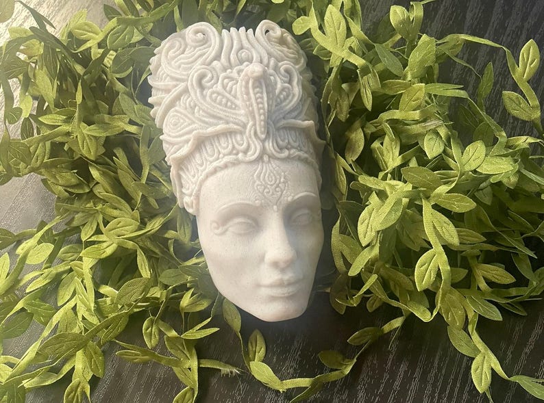 May include: A detailed, off-white sculpture of a regal female bust, adorned with an elaborate headdress featuring a cobra. The bust is surrounded by green leafy vines, creating a naturalistic contrast. The sculpture's face has a serene expression.