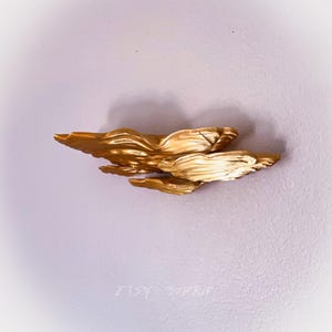 May include: A gold-colored wall sculpture made of wood, with a flowing, organic shape. The sculpture is mounted on a white wall.