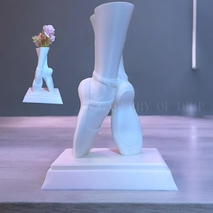 May include: White ceramic ballerina shoe vase with a bouquet of pink flowers. The vase is on a white base.