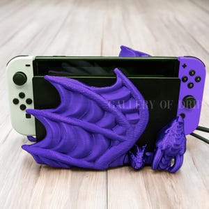 May include: Purple dragon-shaped stand for a Nintendo Switch console. The stand is made of 3D printed plastic and features a detailed dragon design with wings and a head. The Switch console is in the stand, with the Joy-Cons attached.