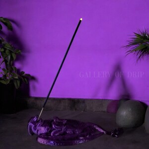 May include: A purple incense holder shaped like a dragon with a black incense stick standing upright in the holder. The holder is on a gray surface in front of a purple wall. The text "GALLERY OF DRIP" is visible on the wall.