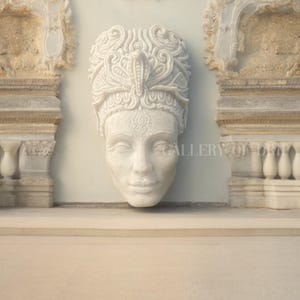 May include: A white marble sculpture of a serene face with ornate headwear, possibly a crown or headdress, is mounted on a light gray wall. The sculpture is framed by architectural elements with intricate carvings. The overall aesthetic suggests classical art.
