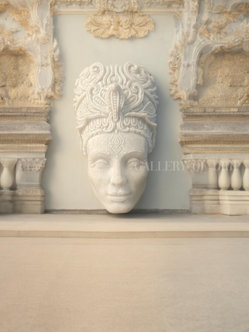 May include: A white marble sculpture of a serene face with ornate headwear, possibly a crown or headdress, is mounted on a light gray wall. The sculpture is framed by architectural elements with intricate carvings. The overall aesthetic suggests classical art.