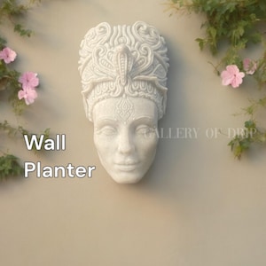 May include: A white wall planter in the shape of a sculpted face with ornate headwear. The planter is mounted on a beige wall, with the words "Wall Planter" in white text. Green vines and pink flowers frame the planter.