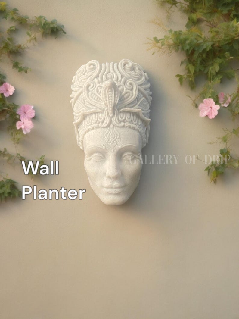 May include: A white wall planter in the shape of a sculpted face with ornate headwear. The planter is mounted on a beige wall, with the words "Wall Planter" in white text. Green vines and pink flowers frame the planter.