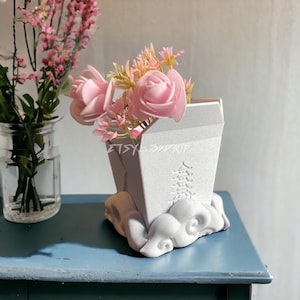 May include: White ceramic planter shaped like a takeout container with a cloud base. The planter has a design of a pagoda on the side. Pink artificial flowers are in the planter.