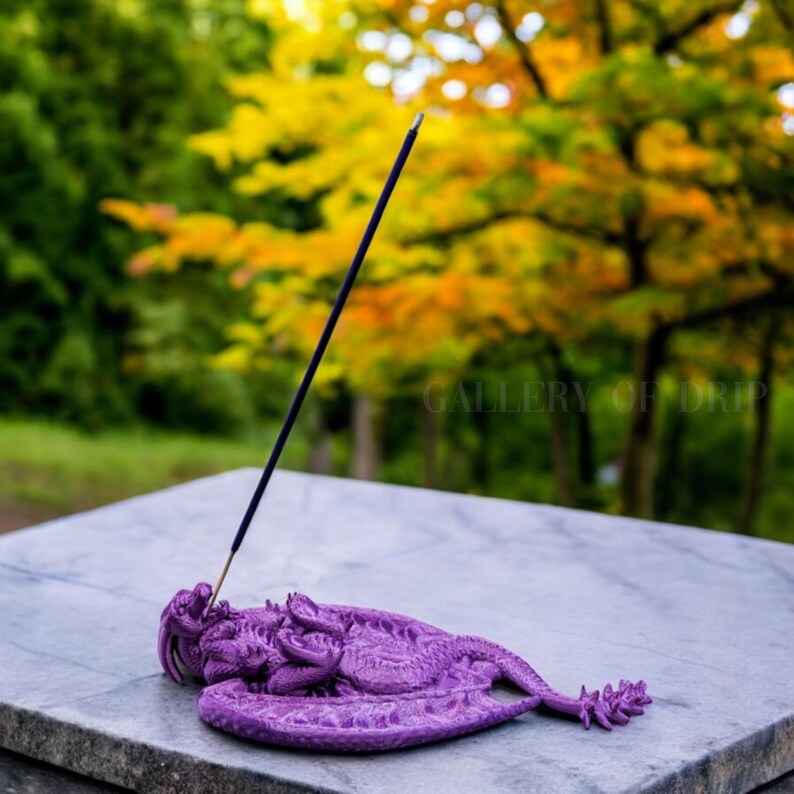 May include: Purple dragon incense holder with a black incense stick standing upright in the holder. The holder is on a white marble surface. The background is blurred and shows green and yellow trees.