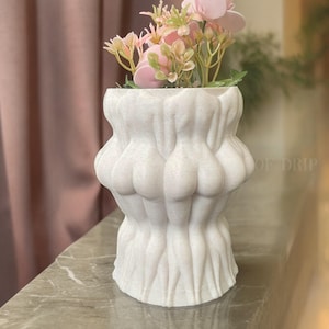 May include: White ceramic planter shaped like a group of figures with pink flowers inside. The planter is on a gray surface.