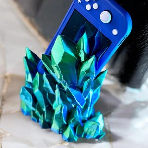 May include: A blue Nintendo Switch Lite console is resting in a 3D printed stand made of iridescent green and blue crystals.