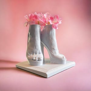 May include: Two white ceramic high-heeled shoes with open toes, used as a vase for a bouquet of pink flowers.