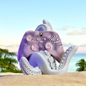 May include: A purple and white swirl Xbox controller is held by a white octopus-shaped stand. The stand has textured tentacles and is on a sandy beach.