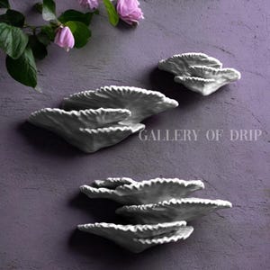 May include: White ceramic wall decor in a layered, organic design. The pieces have a textured, ruffled edge and are mounted on a purple-gray wall. The text "GALLERY OF DRIP" is visible.