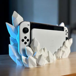 May include: A white Nintendo Switch console rests in a white, crystal-shaped stand. The stand has sharp, geometric facets and is illuminated with blue light. The console's controllers are white and black.