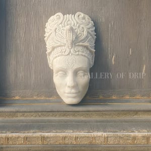 May include: A white, carved stone sculpture of a woman's face with an ornate headdress featuring a cobra. The sculpture is mounted on a gray surface. The text "GALLERY OF DRIP" is visible on the right side.